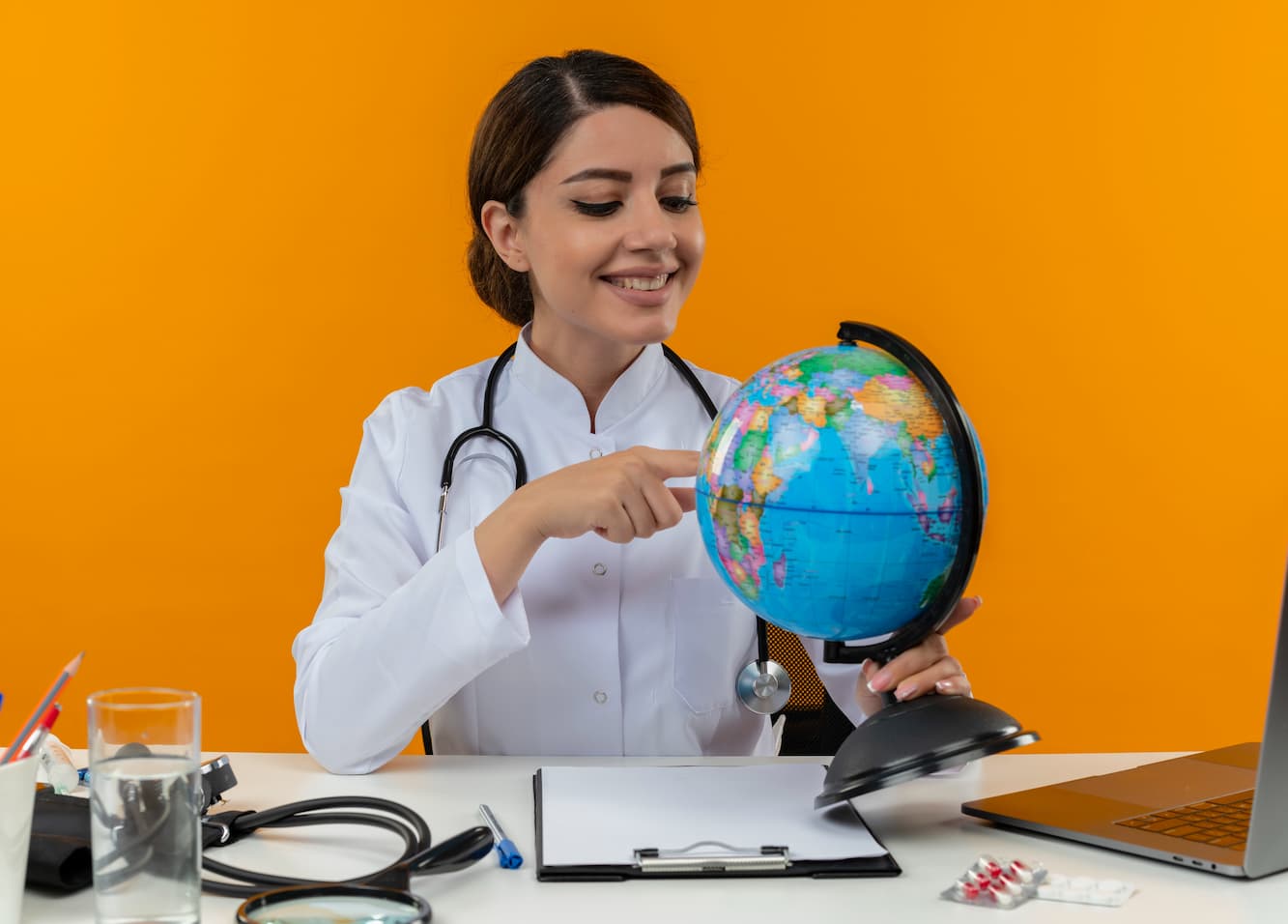 smiling young female doctor wearing medical robe stethoscope sitting desk with medical tools laptop holding looking pointing finger globe isolated yellow wall 1